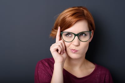 Nerdy scholastic young woman wearing geeky glasses standing thinking with her finger raised and a grimace of concentration in a humorous stereotypical depiction, over a dark background with copyspace